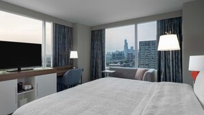 Quarto de hotel com uma cama grande, uma mesa de trabalho e televisão, e vista para o horizonte urbano.