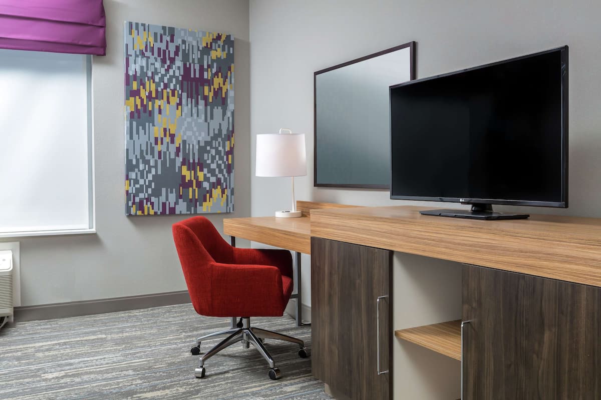 A hotel room with a wooden desk, a red office chair, a flat-screen TV, a wall-mounted lamp, and a colorful abstract painting.