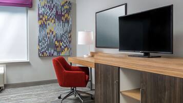A hotel room with a wooden desk, a red office chair, a flat-screen TV, a wall-mounted lamp, and a colorful abstract painting.