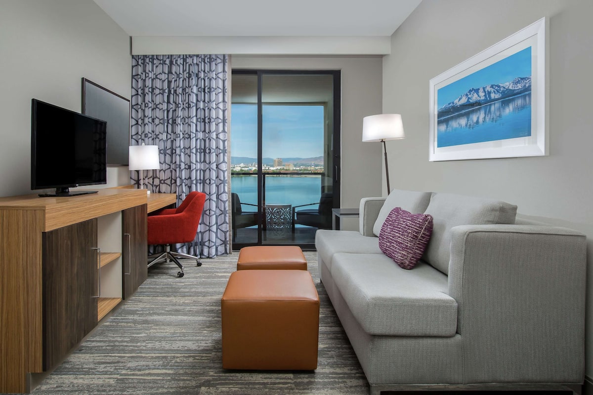 A hotel room with a TV, a red chair, a grey sofa, a purple pillow, and a view of a lake and mountains.