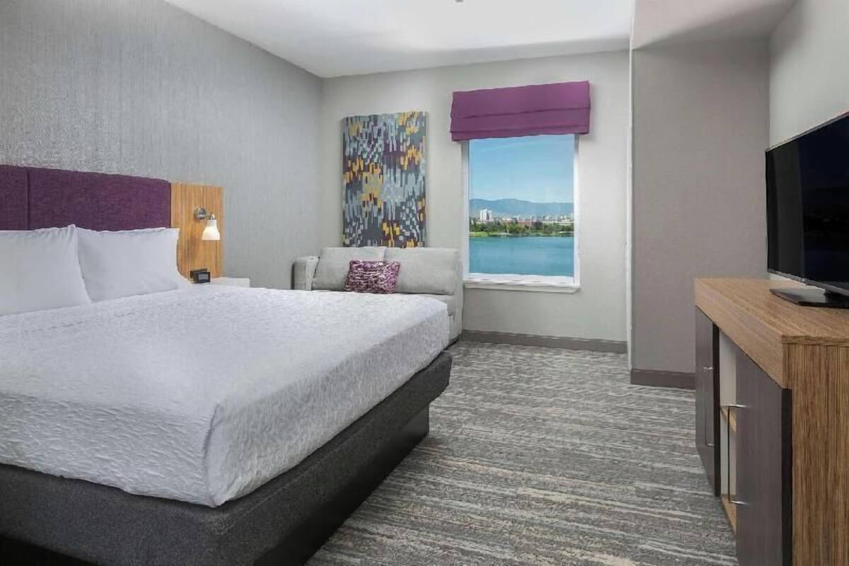 A hotel room with a bed, a TV, a window with a view, a painting, and a lamp.