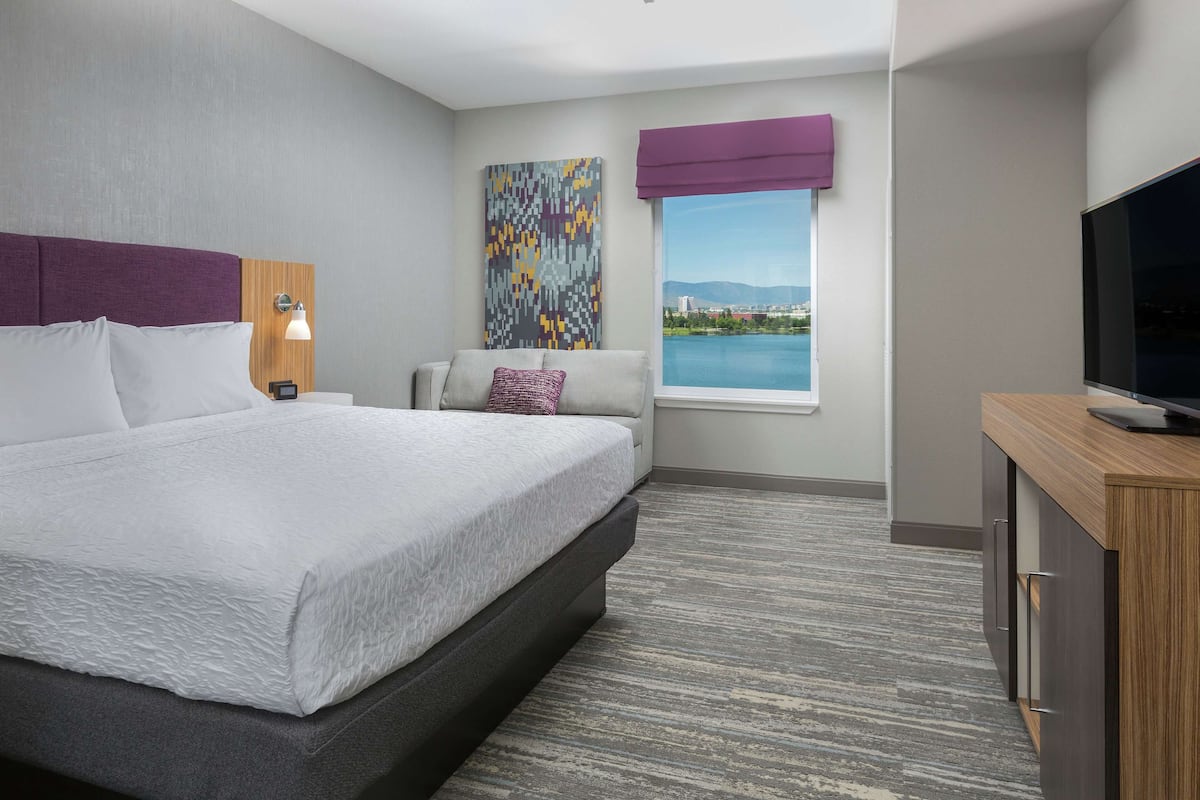 A modern hotel room with a large bed, a television, a desk, and a window with a city view.