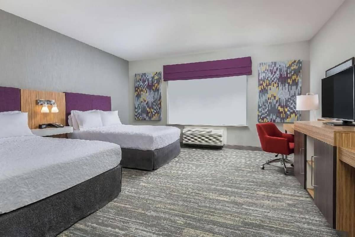A hotel room with two beds, a desk, a TV, and artwork on the walls.