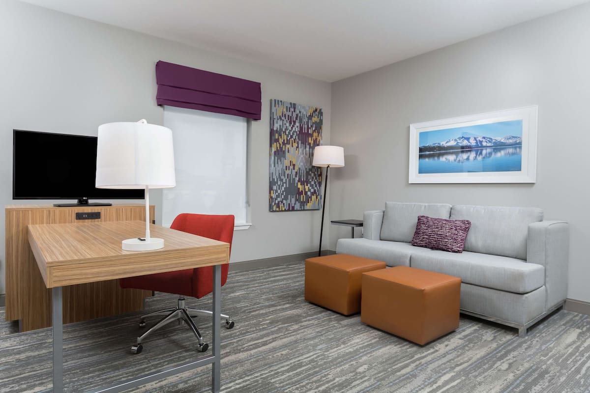 A hotel room with a wooden desk, red chair, grey sofa, two ottomans, a TV, a lamp, a colorful abstract painting, and a framed landscape photo.