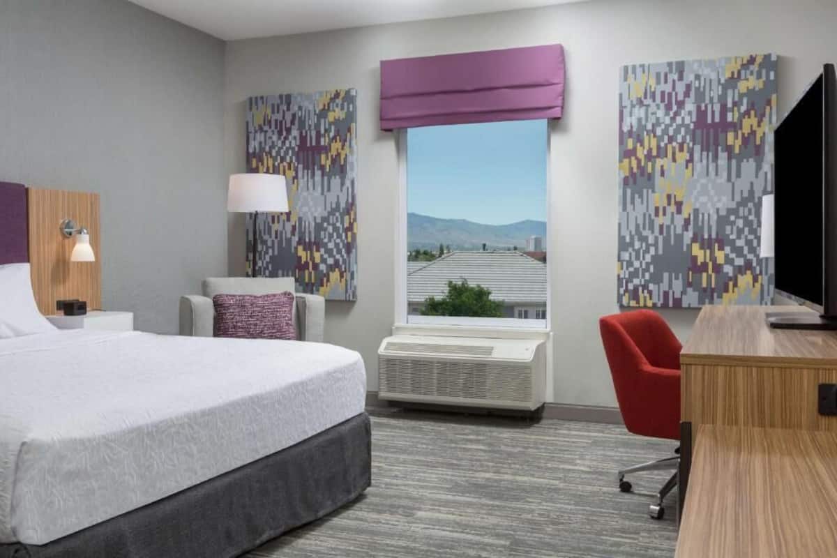 A hotel room with a bed, a desk, a chair, a TV, a window with a view, and artwork on the walls.