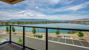 Uitzicht vanaf een balkon op een meer, met bergen op de achtergrond, een parkeerplaats en een heldere lucht.