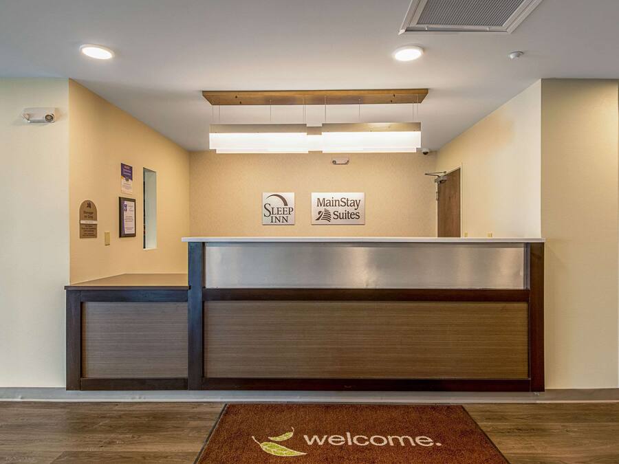 A reception desk with a 'Welcome' mat, 'Sleep Inn' and 'MainStay Suites' signs, and a wooden ceiling.