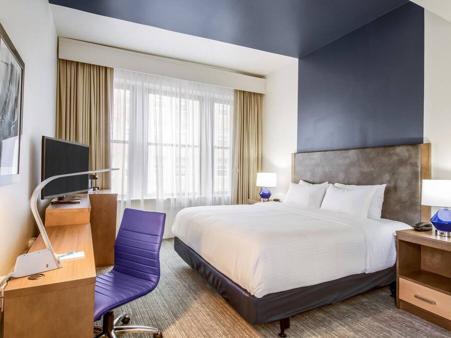 A hotel room with a bed, a desk, a chair, a TV, and a window with curtains.