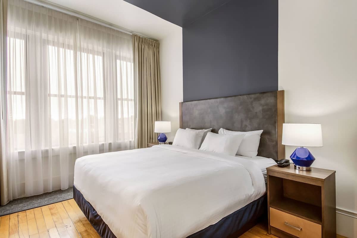 Habitación de hotel con una cama grande, dos mesitas de noche con lámparas, una mesita auxiliar de madera y vistas a la ciudad a través de cortinas translúcidas.