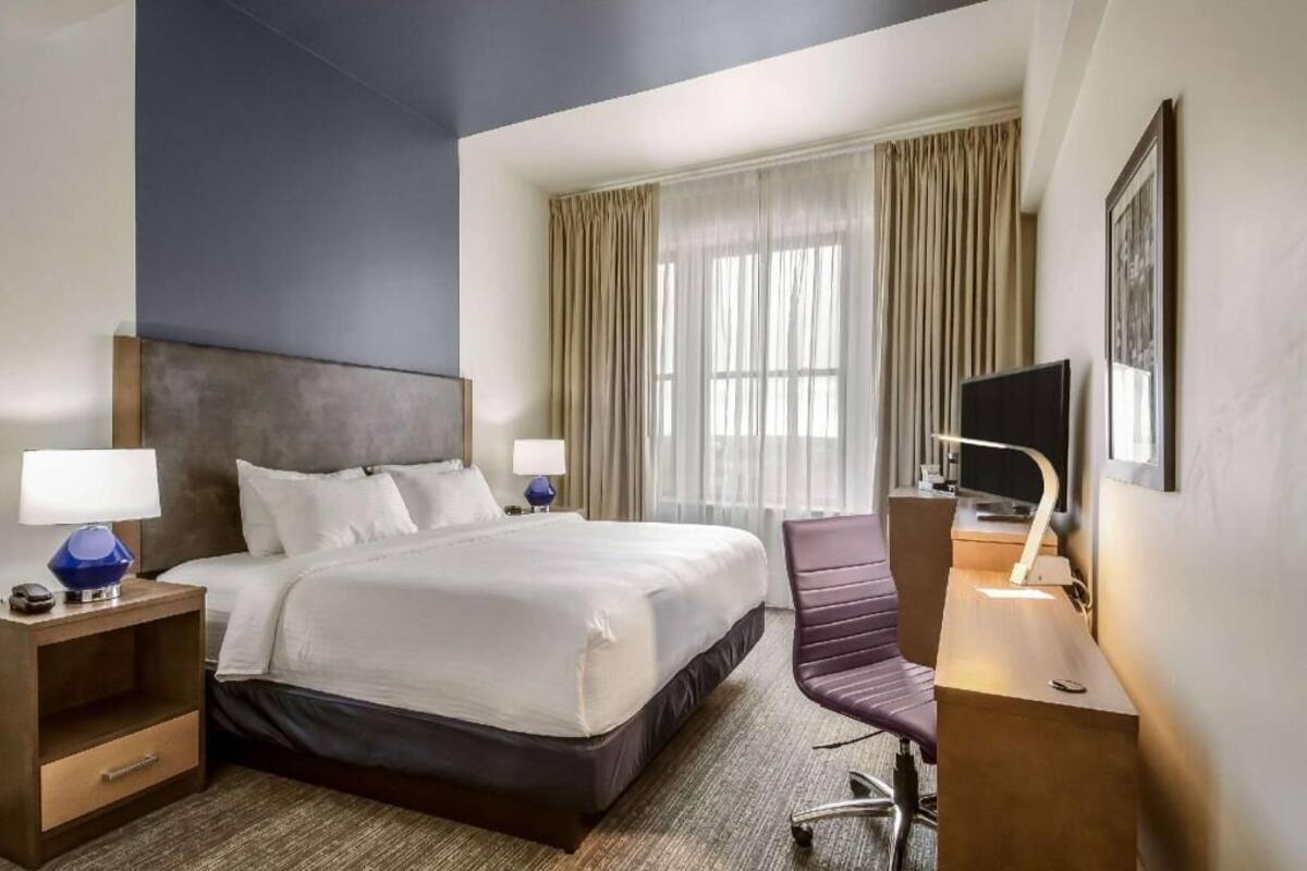 Habitación de hotel con cama, dos mesitas de noche con lámparas, un escritorio con silla, televisor y una ventana grande con cortinas.