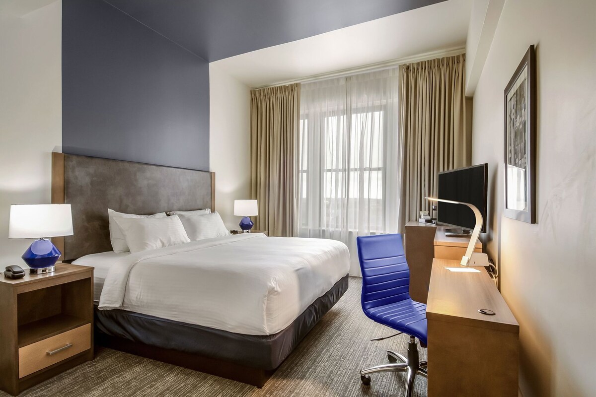 A hotel room with a large bed, two bedside tables with lamps, a desk with a chair, a TV, and a window with curtains.