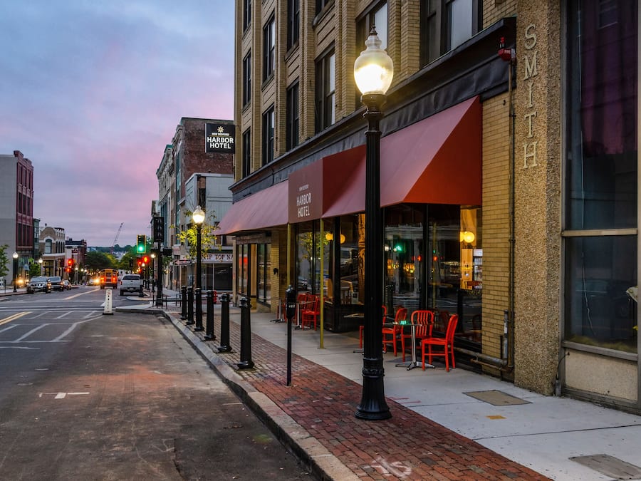 A street view with a hotel named "Harbor Hotel" featuring a red awning and outdoor seating with red chairs.