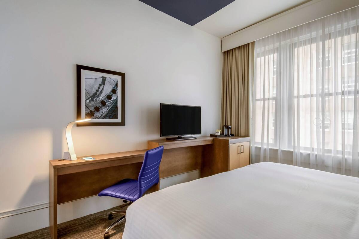 Una habitación de hotel con cama, un escritorio con silla, televisión, un cuadro en la pared y ventana con cortinas.