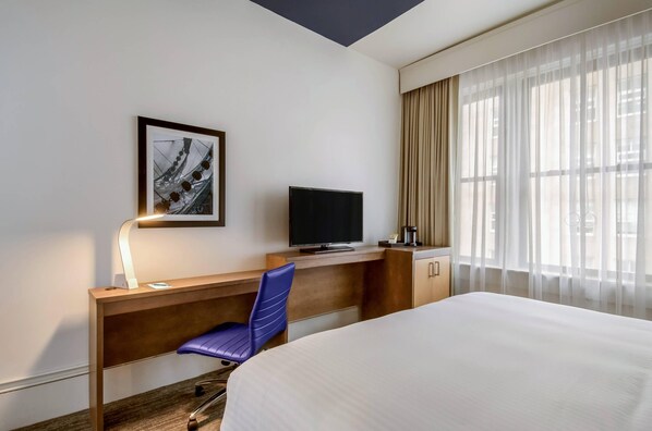 Hotellihuone, jossa on sänky, työpöytä tuolilla, televisio, seinällä oleva taulu ja ikkuna, jossa on verhot.
