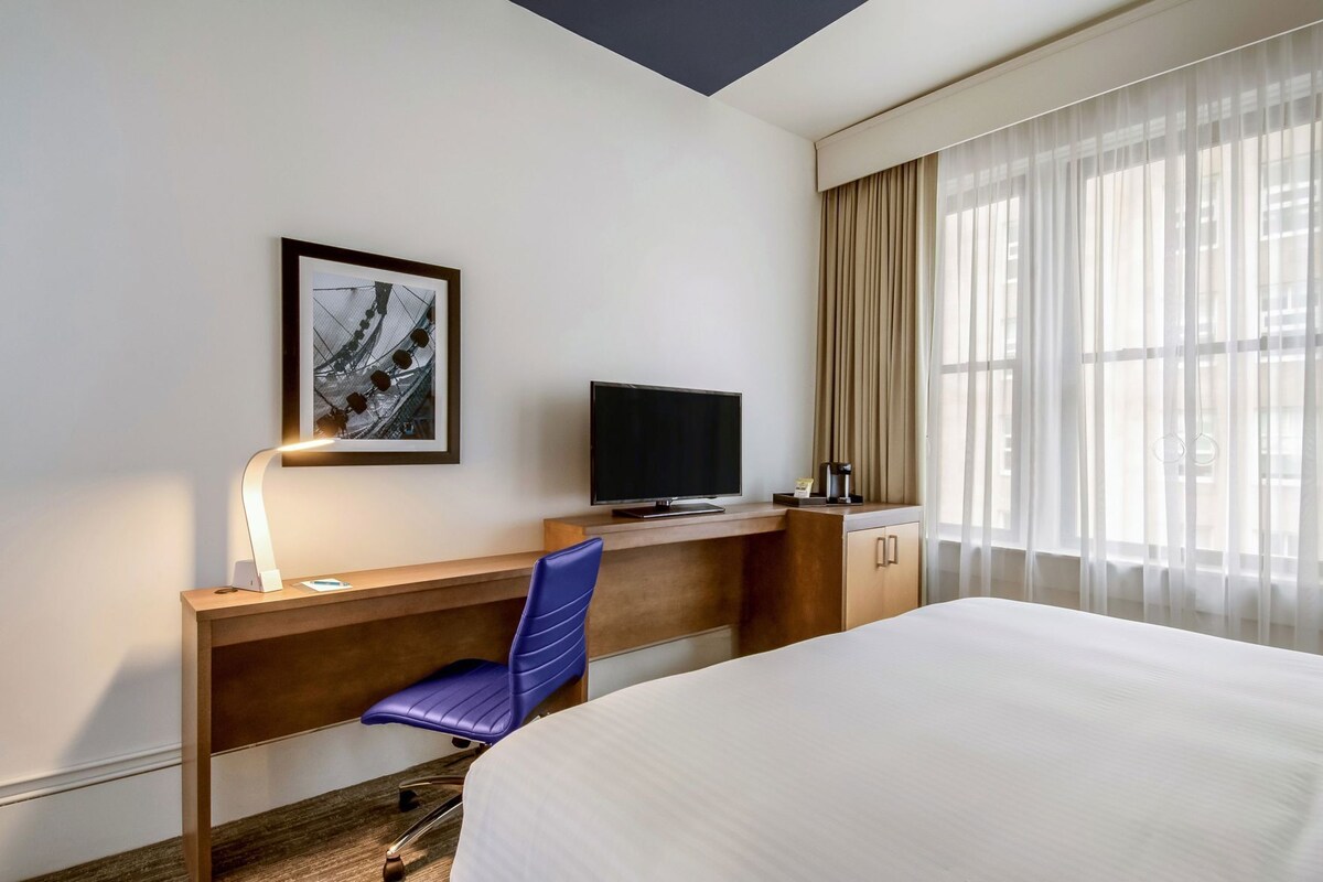 A hotel room with a bed, a desk with a computer, a chair, a TV, and a window with curtains.