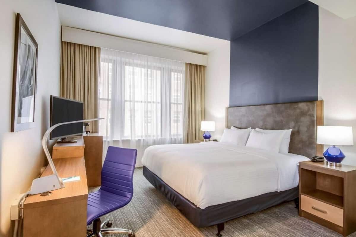 Habitación de hotel con cama, escritorio con computadora, silla, televisor y mesita de noche con lámpara.