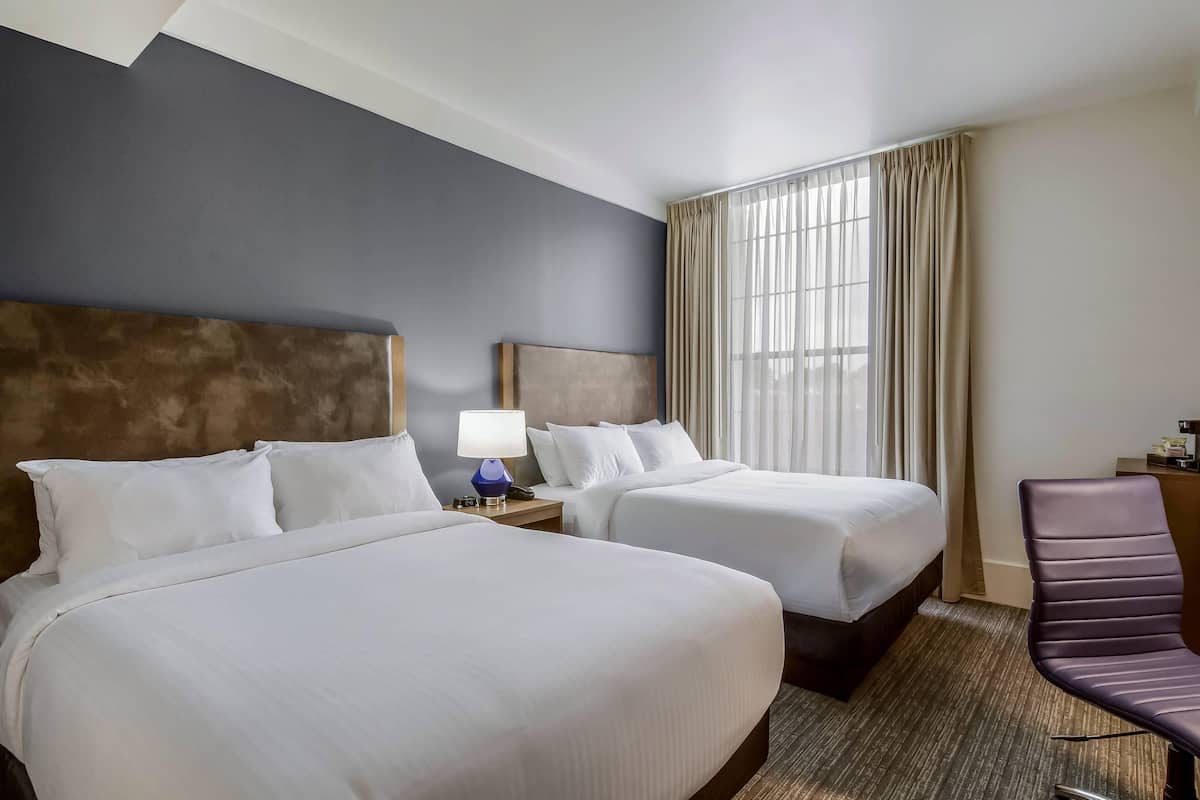 Habitación de hotel con dos camas, una mesita de noche con lámpara, una silla y un ventanal con cortinas.