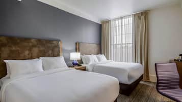A hotel room with two beds, a nightstand with a lamp, a chair, and a large window with curtains.