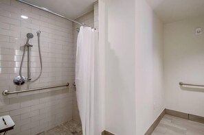 A shower area with a white tiled wall, a showerhead, a curtain, and a handrail.