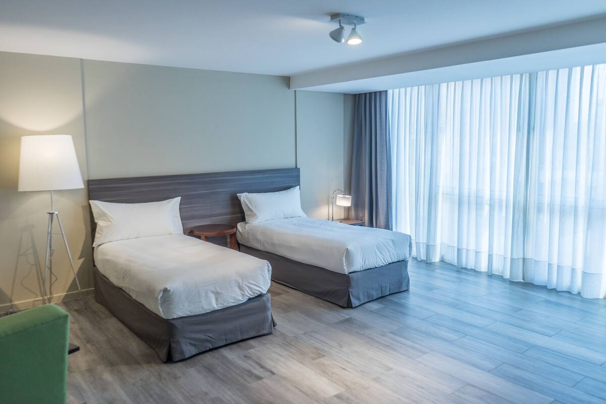 A hotel room with two beds, a standing lamp, and a large window with sheer curtains.