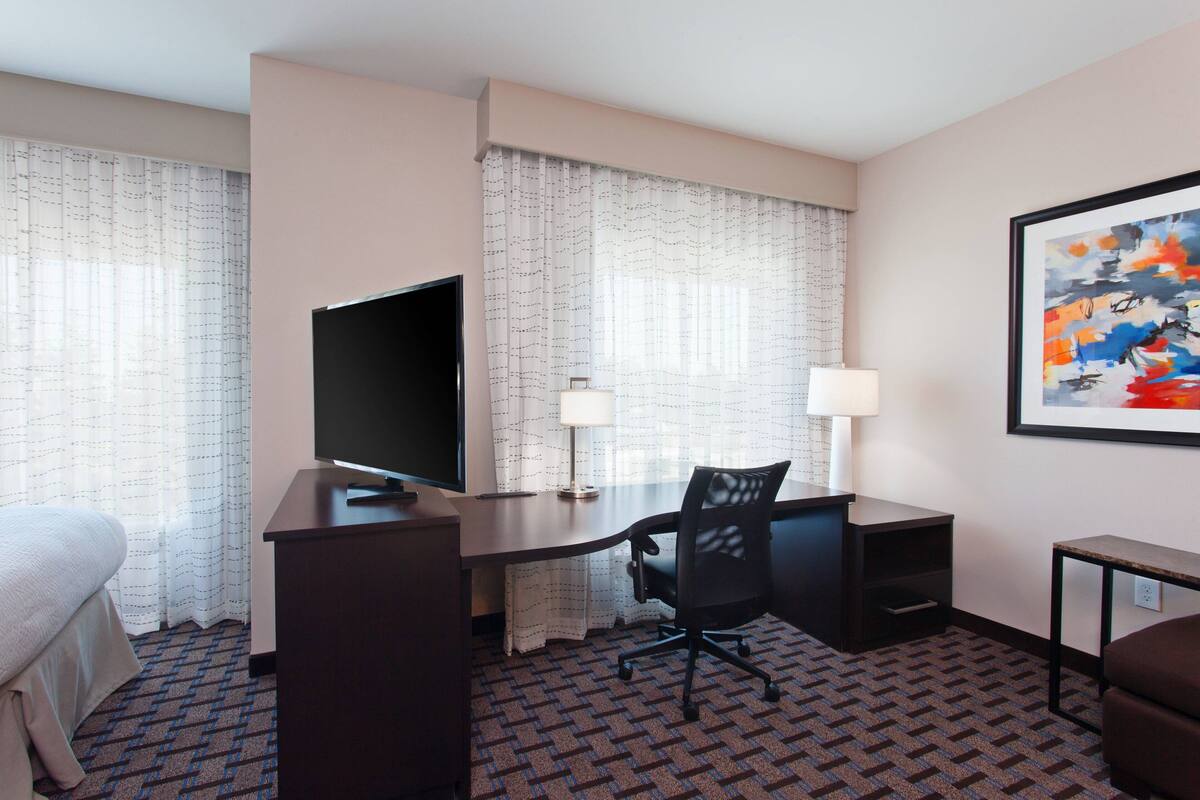 A hotel room with a bed, a desk with a computer, a TV, a lamp, and a painting on the wall.