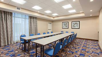 Un salón de conferencias con una larga mesa, sillas azules, alfombra estampada y ventanas grandes con cortinas.