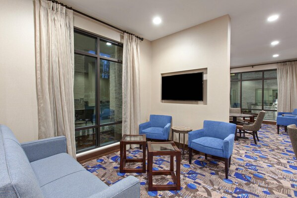 A hotel lobby with a large window, blue armchairs, a small table, and a flat-screen TV mounted on the wall.