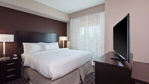 Une chambre d’hôtel avec un grand lit, deux tables de chevet avec des lampes, un téléviseur posé sur un bureau et une fenêtre ornée de rideaux à motifs.