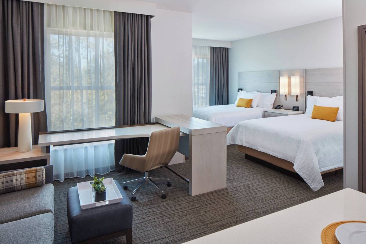 Ein Hotelzimmer mit zwei Betten, einem Schreibtisch, einem Stuhl, einer Lampe und Blick auf Bäume durch das Fenster.