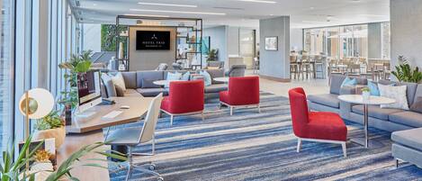 A modern hotel lobby with a seating area, a reception desk, and a dining area.