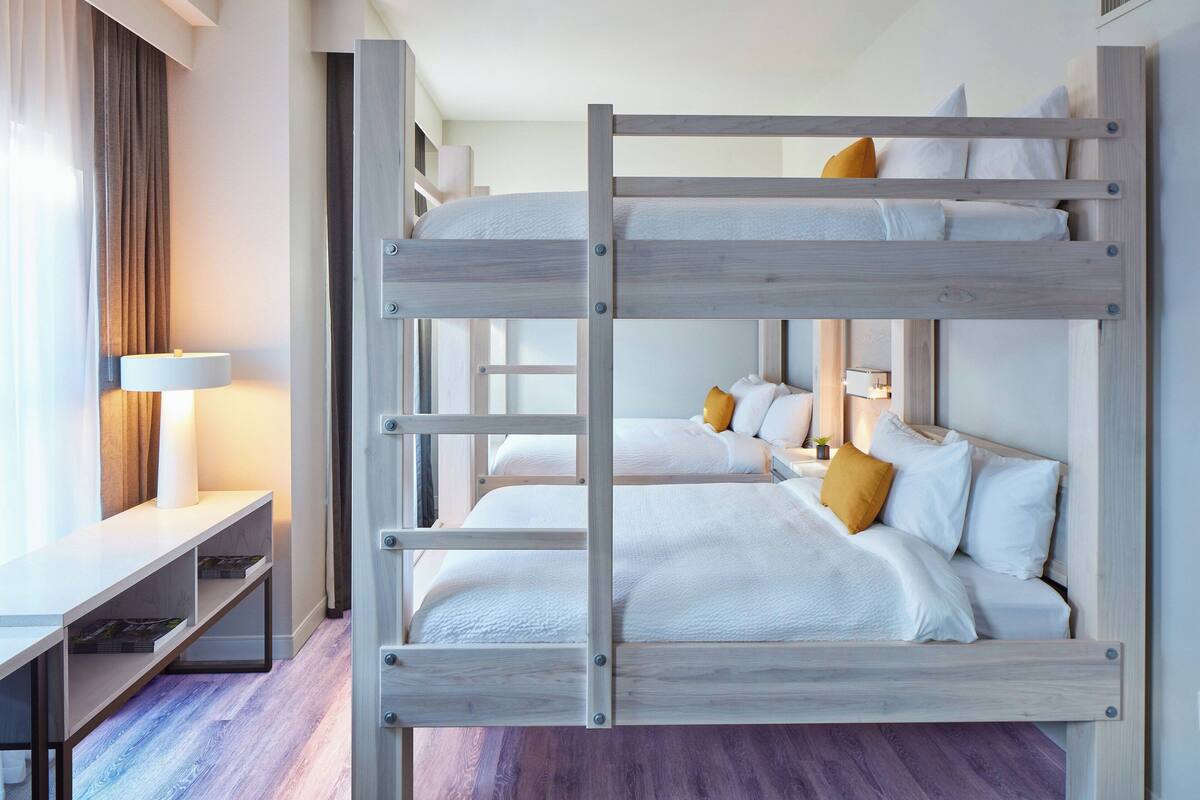 Ein Stockbett mit weißer Bettwäsche und einem Holzrahmen, ein Schreibtisch mit Lampe und ein Fenster mit Vorhängen.