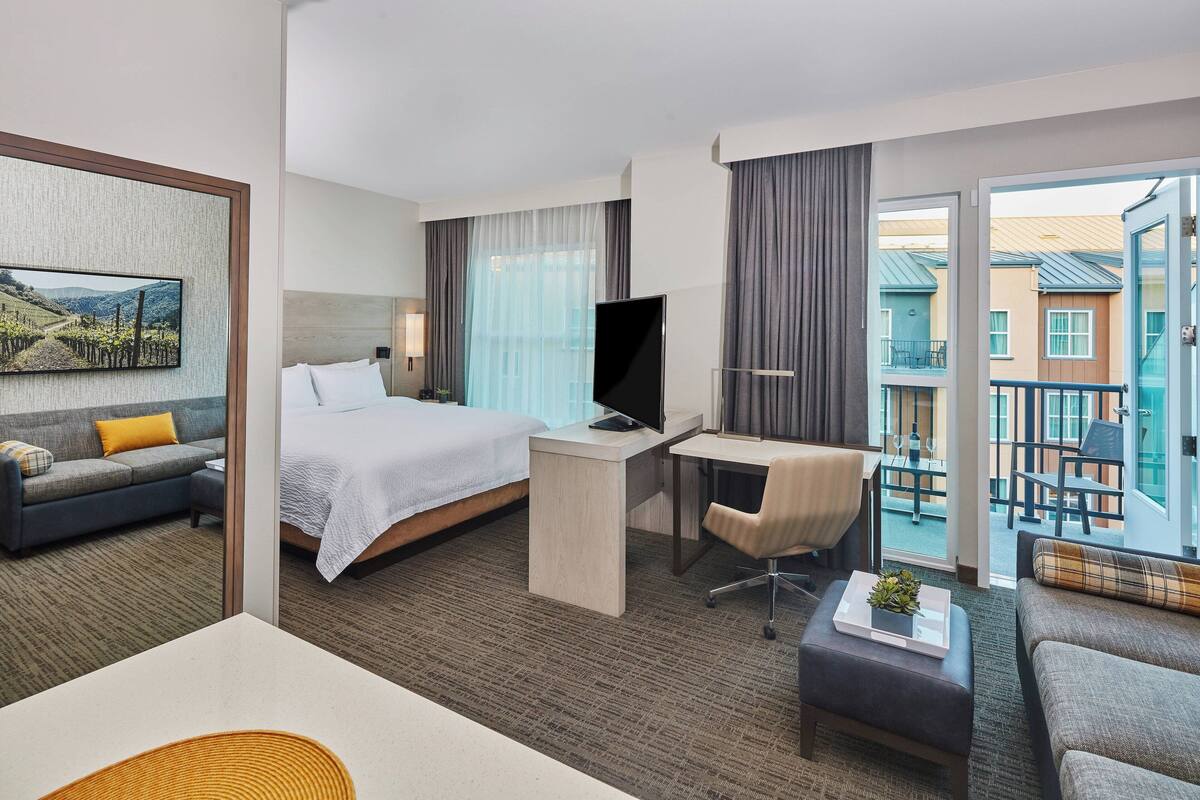 Ein Hotelzimmer mit Bett, Schreibtisch, Fernseher und einem Balkon mit Blick auf Gebäude.