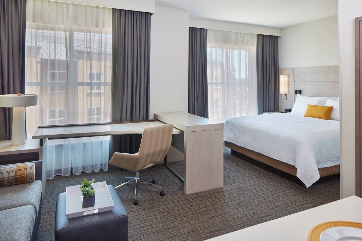 Ein Hotelzimmer mit Bett, Schreibtisch, Stuhl, einem kleinen Tisch mit Pflanze und einem Fenster mit Vorhängen.