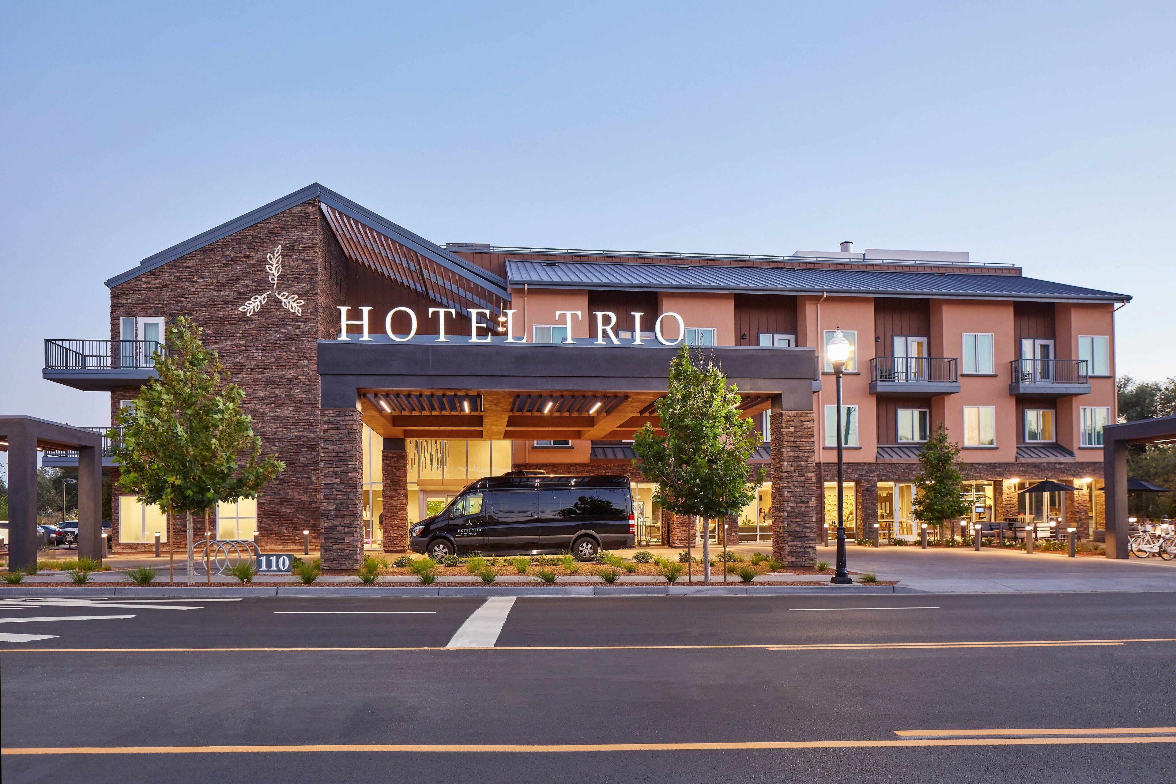 A hotel named HOTEL TRIO with a modern design, featuring a black van parked in front, a well-lit entrance, and a clear view of the building's facade.