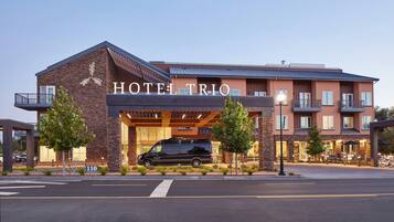 A hotel named HOTEL TRIO with a modern design, featuring a black van parked in front, a well-lit entrance, and a clear view of the building's facade.
