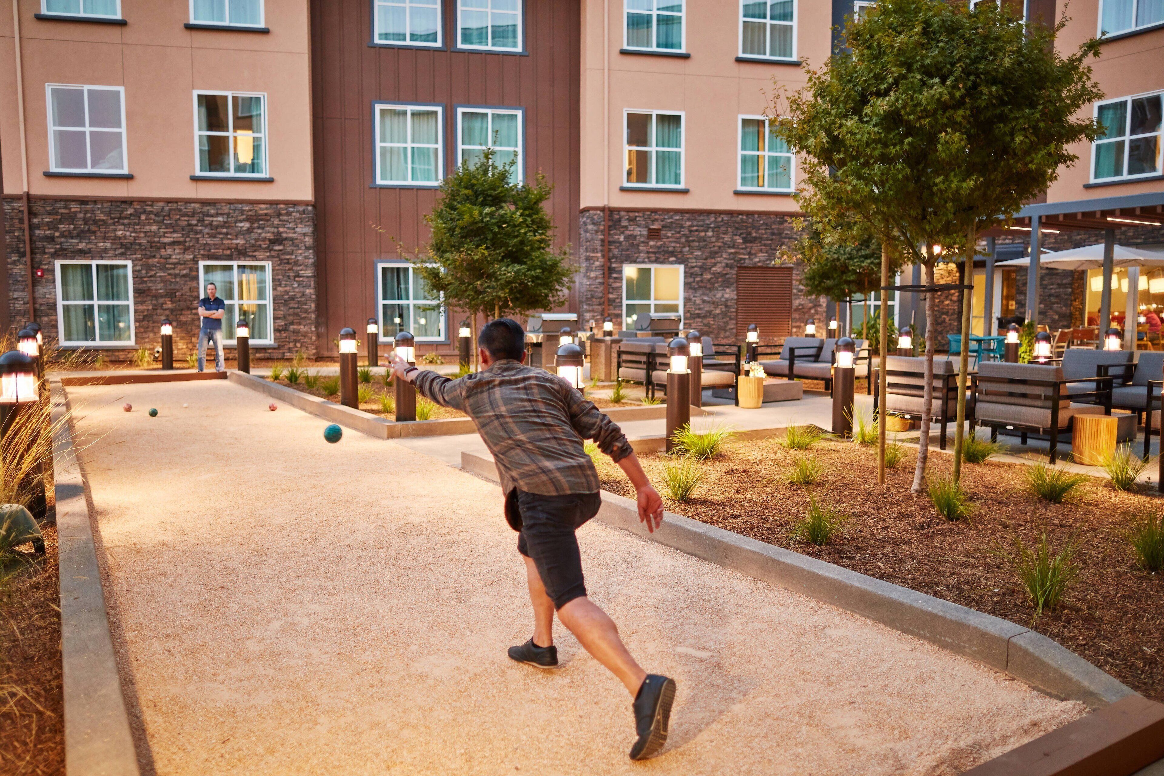 A person playing shuffleboard in an outdoor area with a building in the background.