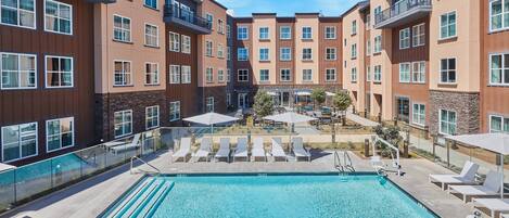 A modern apartment building with a central courtyard featuring a swimming pool, lounge chairs, and outdoor umbrellas.