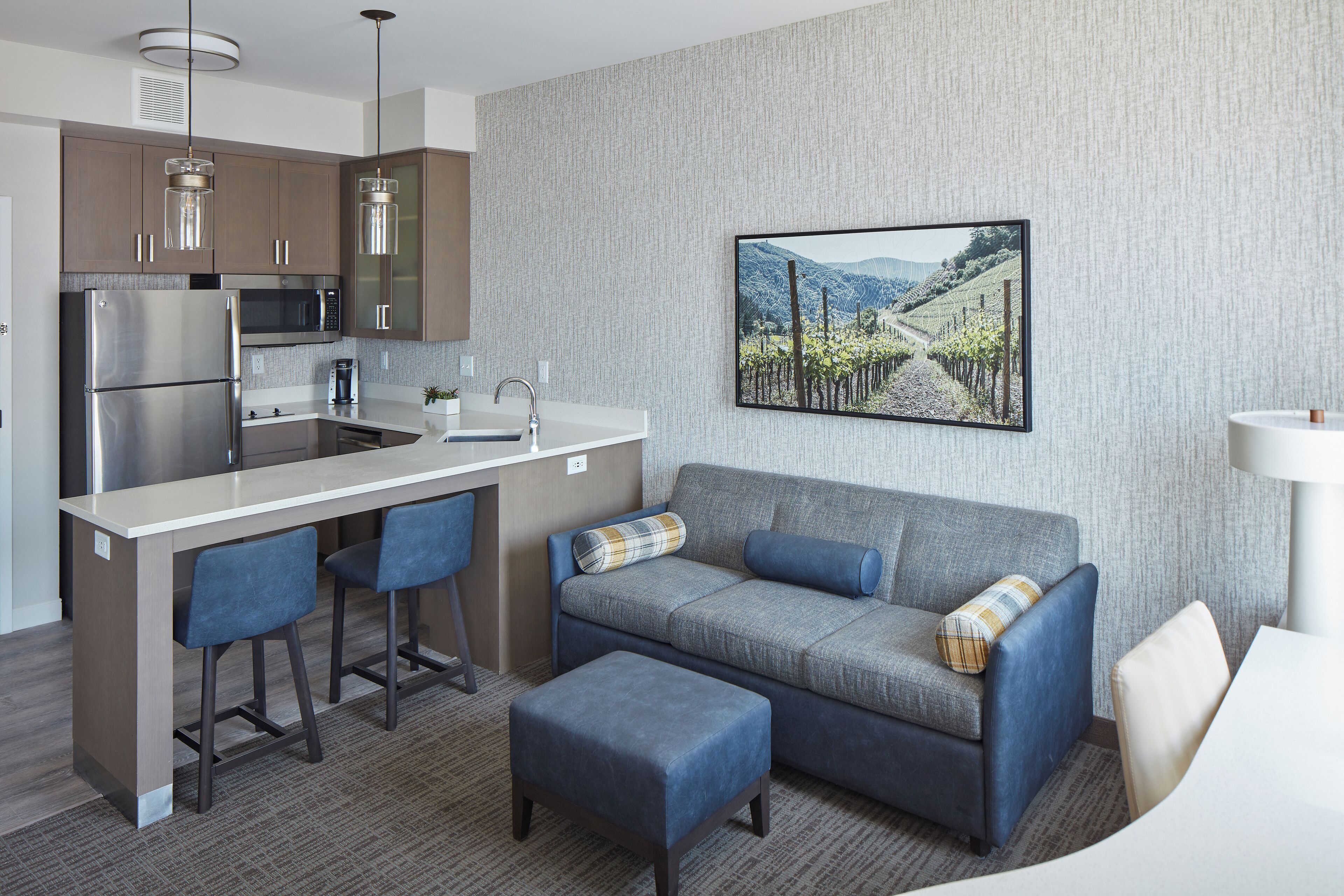 A modern kitchen with a built-in refrigerator, microwave, and coffee maker. There is a bar area with stools and a gray sofa with blue cushions. A framed picture of a vineyard is on the wall.