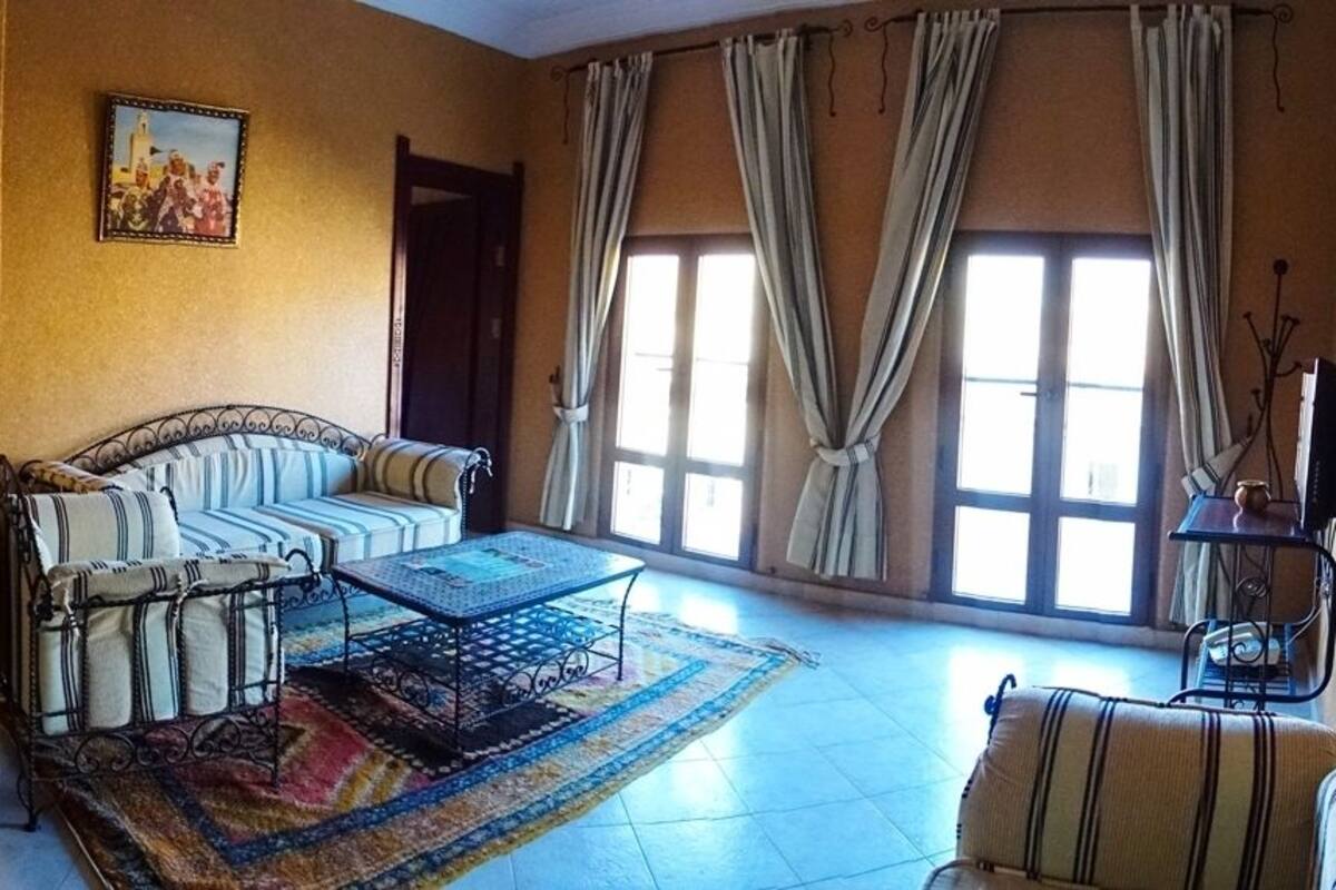 Un salón con un sofá, una mesa de centro y una silla. Hay una ventana con cortinas y un cuadro en la pared.