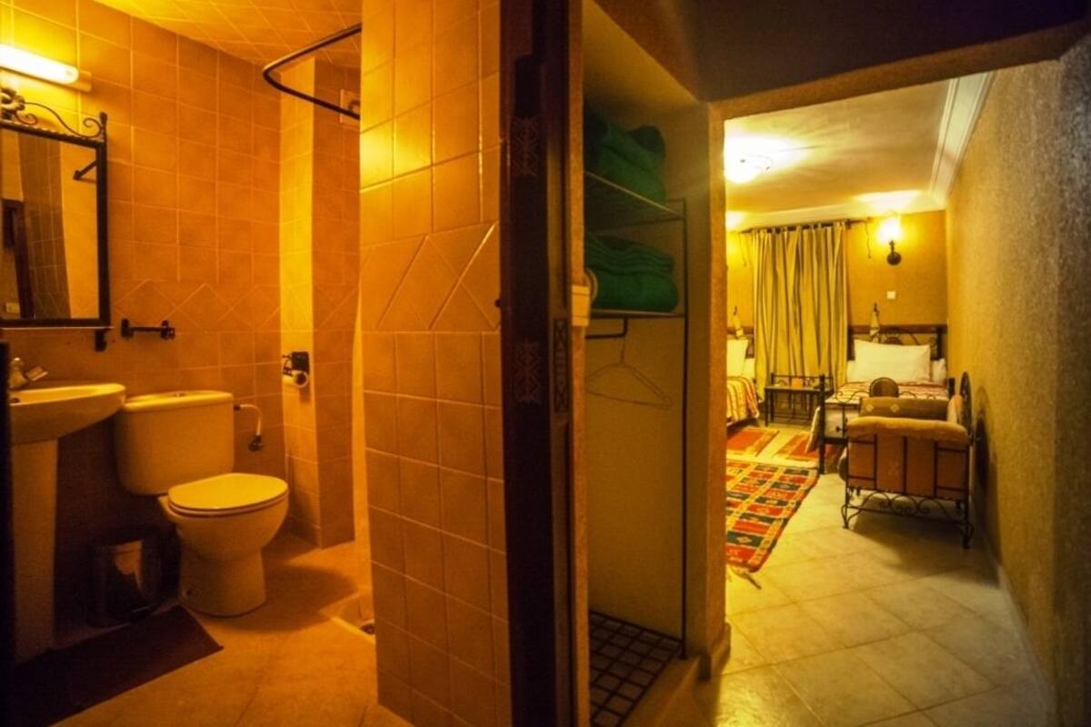 Un baño de hotel con ducha, inodoro y lavamanos. Se ve un pasillo que conduce a un dormitorio con cama y mesita de noche.