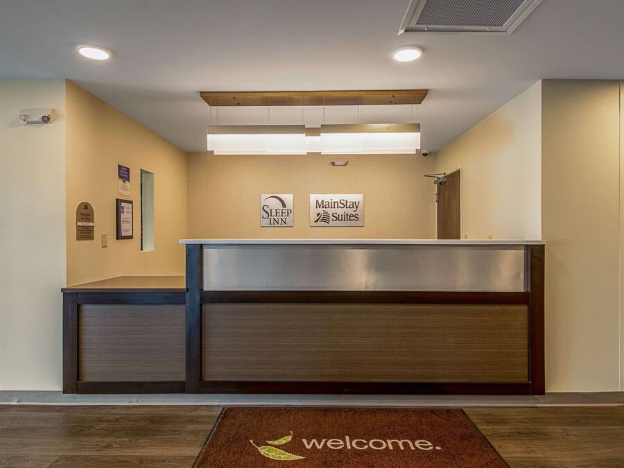 A reception desk with a 'Welcome' mat, 'Sleep Inn' and 'MainStay Suites' signs, and a wooden ceiling panel.