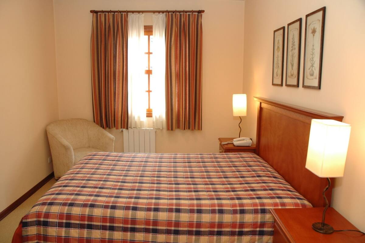 A hotel room with a bed, a chair, a bedside table with a lamp, a window with curtains, and framed pictures on the wall.