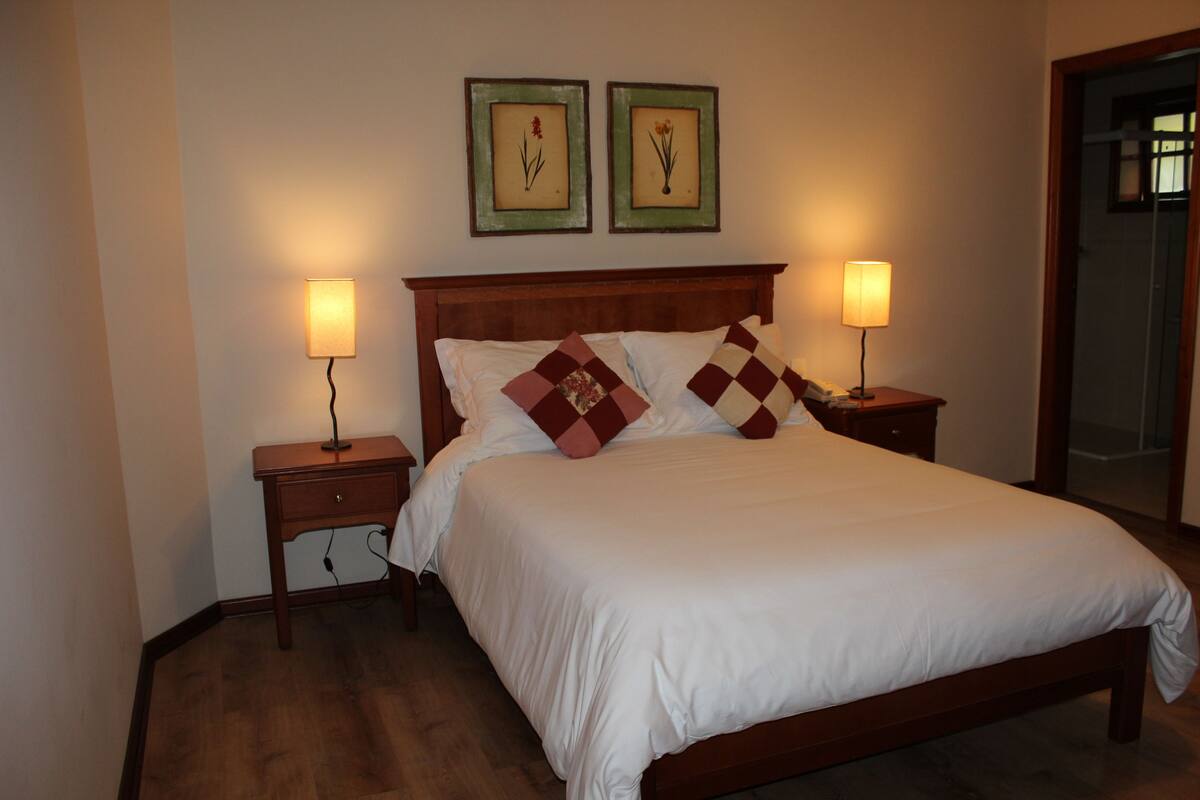 A neatly made bed with white bedding and patterned pillows, flanked by two bedside tables with lamps, in a room with wooden flooring and a wooden headboard.