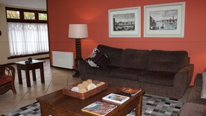 A living room with a brown sofa, a coffee table with books and a basket, a lamp, and two framed pictures on the wall. - Pousada Chablis (Campos do Jordão)