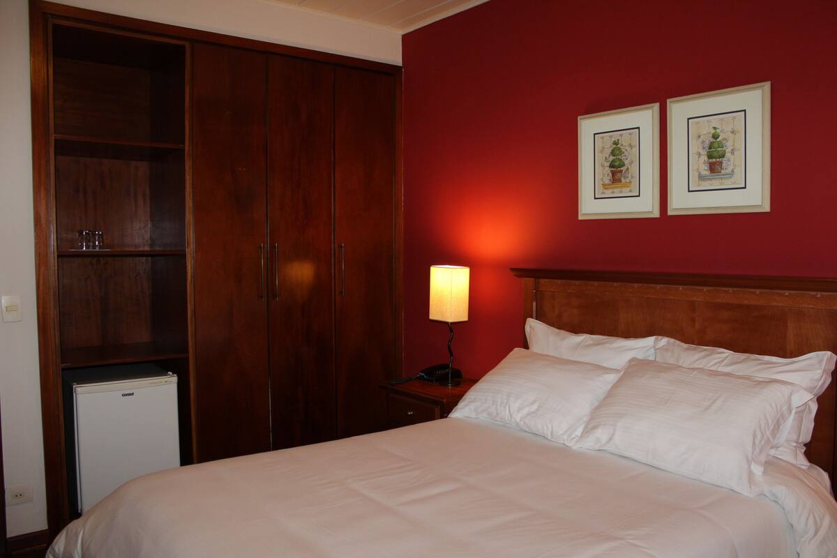 A hotel room with a bed, a wardrobe, a nightstand with a lamp, and two framed pictures on the wall.