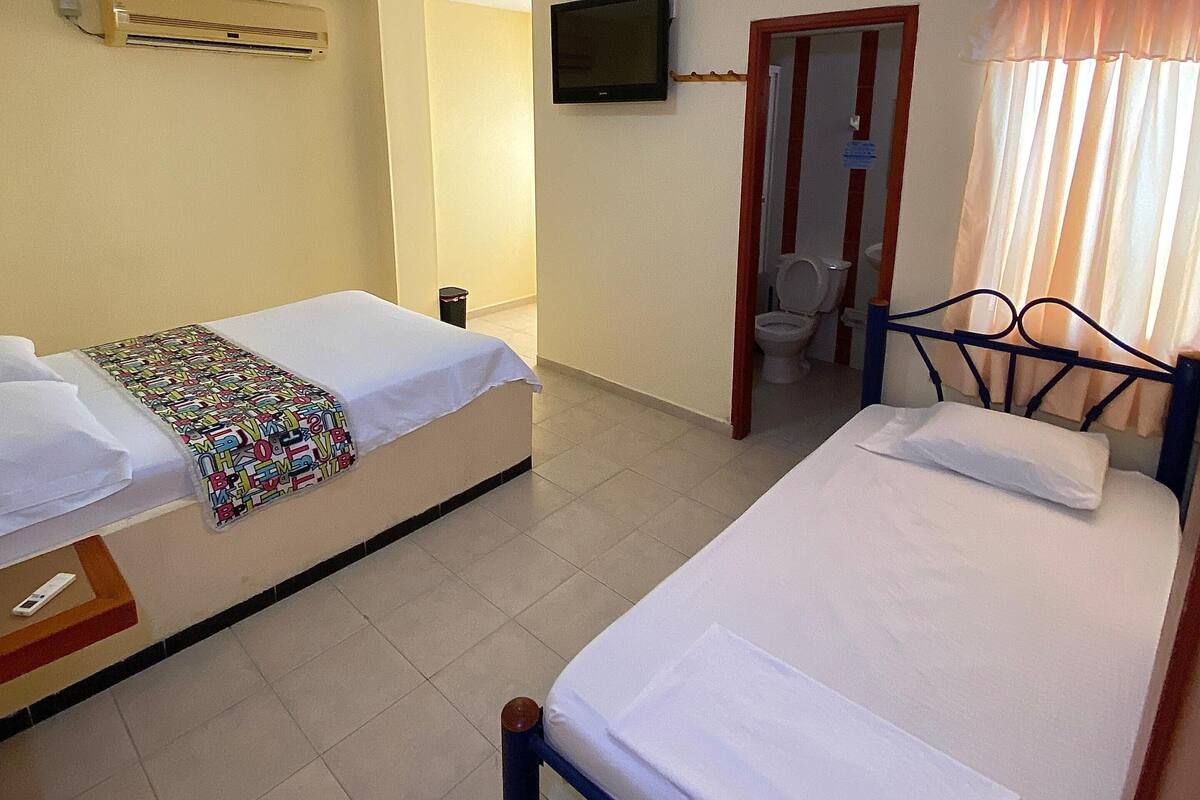Habitación de hotel con dos camas individuales, un televisor fijado a la pared, baño con inodoro y lavamanos, y ventana con cortinas.