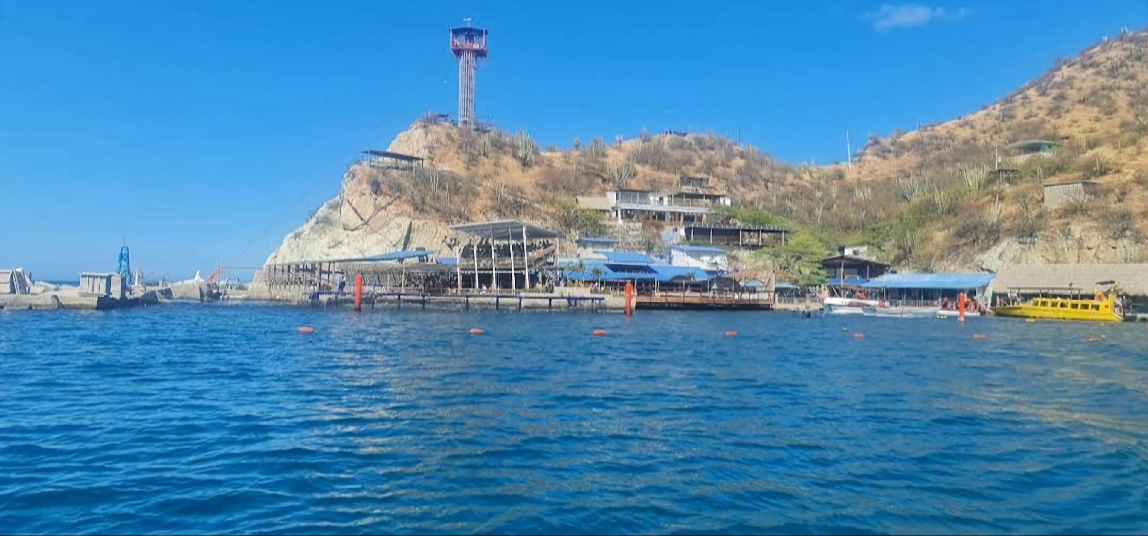 A coastal area with a lighthouse, a pier with covered structures, and boats on the water.