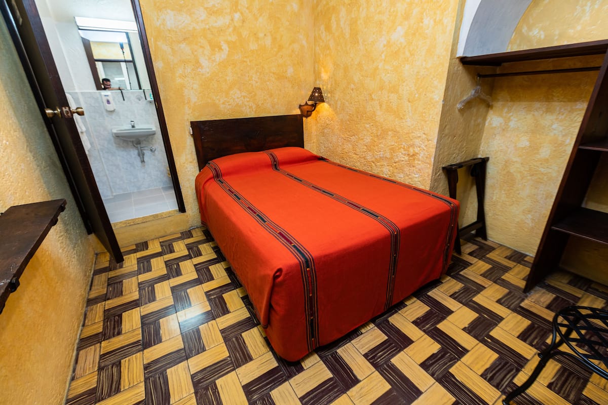 A room with a bed, a wooden floor, a wall-mounted light, and a bathroom visible through an open door.