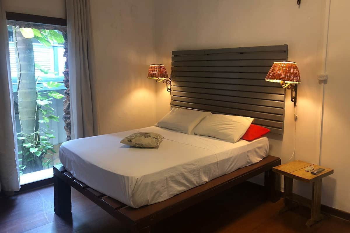 A bed with a wooden headboard, two bedside lamps, a bedside table with a phone, and a view of greenery through a window.