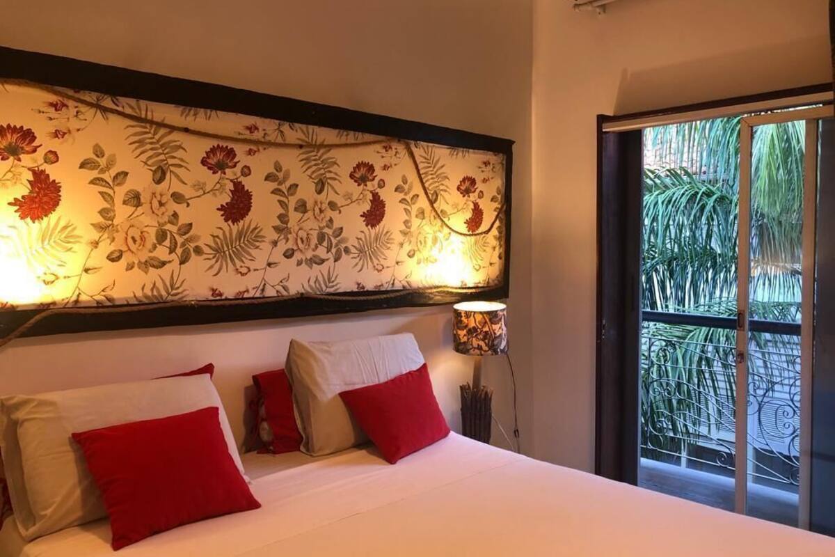 A hotel room with a bed, red pillows, a floral headboard, a lamp, and a view of greenery through a sliding door.